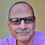 man with buzz cut and mustache wearing sunglasses and purple t shirt smiles in front of brick wall