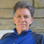 man with short grey hair and blue jacket sitting in chair poses for photo
