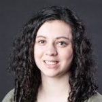 A smiling person with curly hair is facing the camera against a dark background.