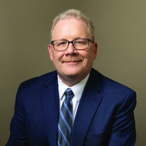 A headshot of Willy McKain, in a suit and glasses, smiling