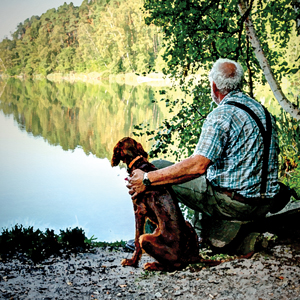 A man sitting by a lake with a dog, you can see the reflection of the trees in the lakes water