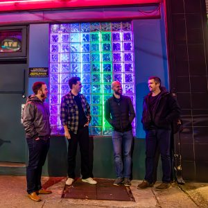 Four men standing on sidewalk with neon lights behind them