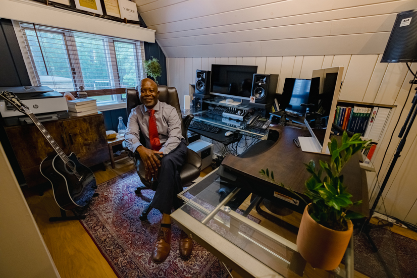 a man sitting in a chair at his desk in his home office, there is an acoustic guitar in the corner and several screens, speakers, and electronic equipment on the desk