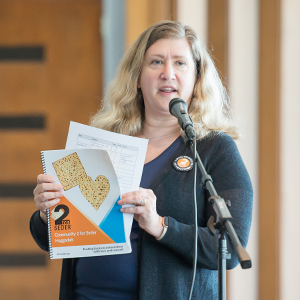 Woman with blonde hair wearing a navy blue sweater and top standing in front of a microphone holding the 2 for Seder event program book.