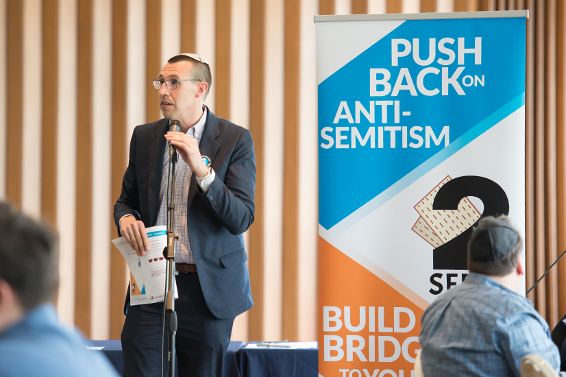 A man with very short black hair wearing a white yamaka, glasses and a blue plaid button down shirt under a navy blue blazer standing in front of a microphone stand holding the 2 for Seder event program with a pull up banner in the background with white text over a bright blue background that says "push back on anti-semitism" at the top and white text over an orange background that says "build bridges" on the bottom.