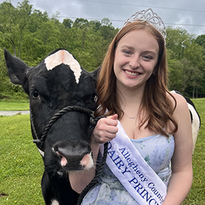 Young lady with light brown hair wearing a formal pastel dress with flowers, a white sash with blue text that says Allegheny County Dairy Princess, and a sparkly silver tiara and her black and white cow in front of vibrant green trees.
