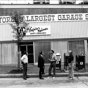 A black and white photo of several men standing in front of a building, a sign says "the worlds largest garage sale"