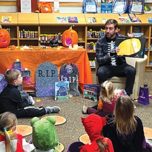 A man in a halloween costume reading a book to children also in halloween costumes. The library is decorated for halloween with tomb stones and jack-o-lanterns