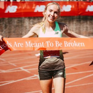 A woman at the running finish line holding up a ribbon with a quote on it that says "Records are meant to be broken"