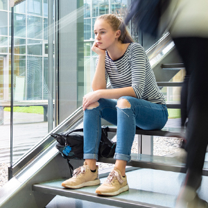 Teen girl in a black and white striped shirt, ripped jeans and sneakers sitting on a set of stairs looking sad with a blurry person wearing a white shirt and black pants carrying a backpack in the foreground.