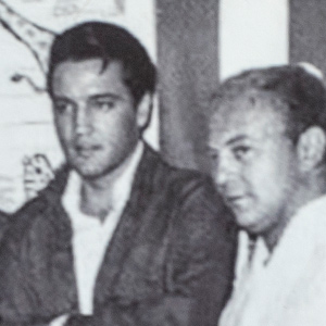 Black and white vintage photo of Elvis Presley (wearing a collared shirt and a dark coat) with a man standing to the right.