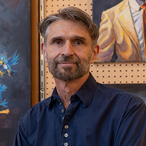 A man with short light brown hair and beard wearing a navy blue button down collard shirt standing with arms crossed.