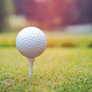 Close up photo of a white golf ball sitting on a white tee with green grass and greenery blurry in the background.