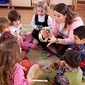 A group of preschool students sitting in a circle on a rug playing with musical instruments with their teacher showing them how to use a tambourine.