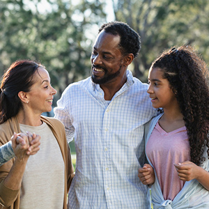 A multiracial family standing together outdoors, trees in the background. The parents are standing to the left with their daughter on the right.