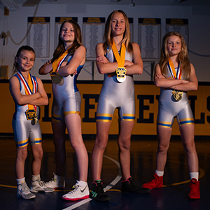 A group of 4 girls smiling and wearing silver wrestling uniforms and multiple medals on ribbons around their necks standing in front of a wall with the lower section painted yellow with the words Blue Devils in navy blue.