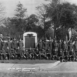 A black and white photo of a lot of men dressed in uniform lined up in front of a memorial