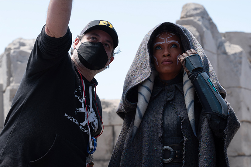 Man wearing a black baseball hat, a black face mask and a black sweatshirt with the sleeve pushed up to his elbow pointing up standing by a person with a white tribal patterned face paint dressed in a light gray hooded coat with a plastic piece over the arm with her hand by her face.
