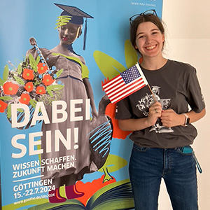 Young female with dark brown hair pulled back with black glasses sitting on top of her head wearing jeans and a dark gray t-shirt with a white graphic on it holding a small American flag standing in front of a light blue banner with a statue wearing a graduation cap holding an illustrated bouquet of bright red flowers with white German words over top.