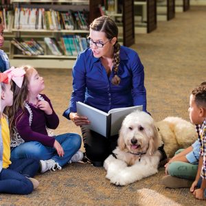 kids sitting on a floor listening to an adult read a book next to a dog