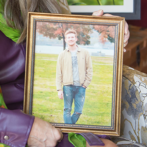 Close up of a woman's hands holding a gold framed photo of her son standing on the grass with large trees with red leaves in the background.
