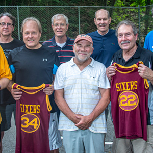 Several men standing in an outside basketball court, two men are holding up Sixers jerseys.
