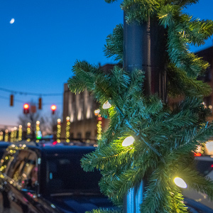 Close up of a light pole wrapped in evergreen garland with bright with lights on Washington Road streetscape at night with parked cars and a traffic light in the background.
