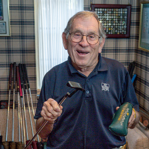 A man with short gray hair and glasses wearing a navy blue short sleeved golf shirt smiling and holding a putter and a forest green putter cover standing in front of golf memorabilia and a wall with a window and plaid wallpaper.
