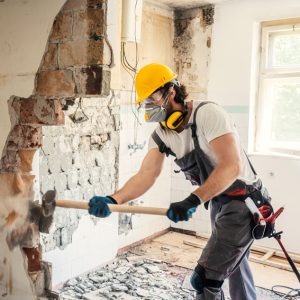 man with sledgehammer and short sleeve t-shirt breaking down a wall, wearing a helmet and mask