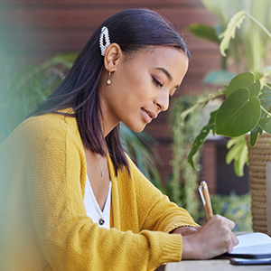 Profile photo of a young lady with dark brown hair wearing a mustard yellow cardigan over a white shirt at a desk writing in a journal surrounded by leafy green plants.