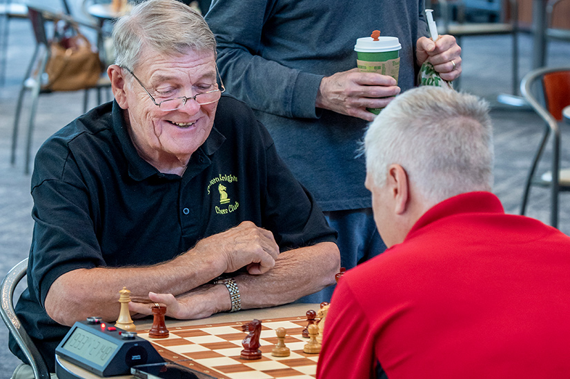 Two men both with short grey hair sitting at a table playing a game of chess. The man facing the camera is wearing a black golf shirt with the words Southern Knights Chess Club embroidered in yellow. The man in the foreground is wearing a bright red golf shirt.