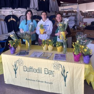two teenage girls and a woman at a table, selling daffodils. table has yellow tablecloth with American Cancer Society written on it.