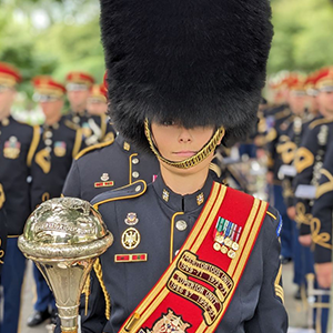 A member of a military band stands at the forefront, wearing a tall black bearskin hat and a dark blue uniform adorned with gold trim, medals, and a red sash. Band members in matching uniforms and red-trimmed hats stand in formation, holding brass and percussion instruments in an outdoor space with green trees in the background.
