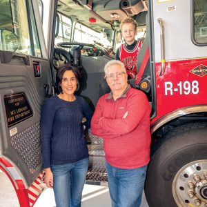 A man and woman standing in front of a fire truck with the front door open, their son a young boy, is sitting in the drivers seat of the firetruck.