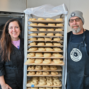 Man and woman smiling next to a rack of bagels