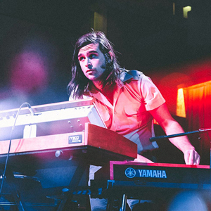 A man with long hair standing on a stage behind several keyboards playing music.