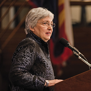 A women with short gray hair speaks into a microphone at a podium, wearing a dark patterned jacket. Blurry flags and a railing are visible in the background.