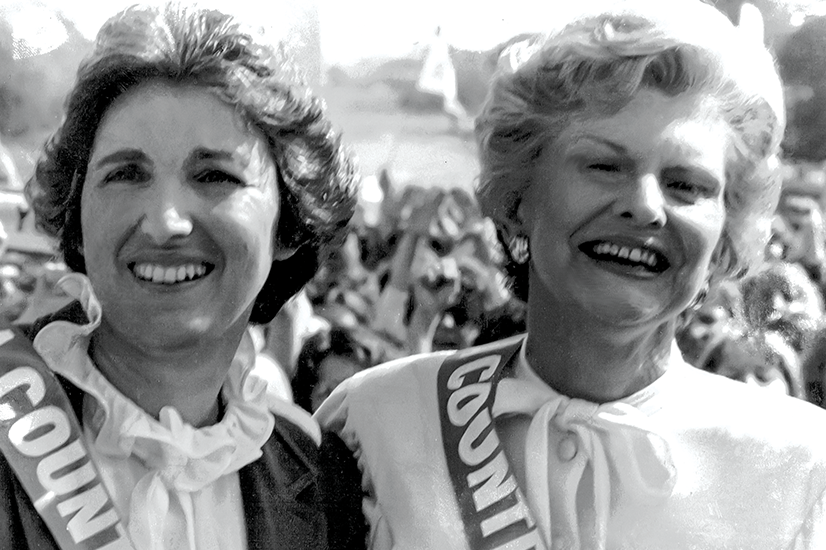 A black and white photo of two women with short hair standing together smiling and wearing sashes in front of a large crowd.