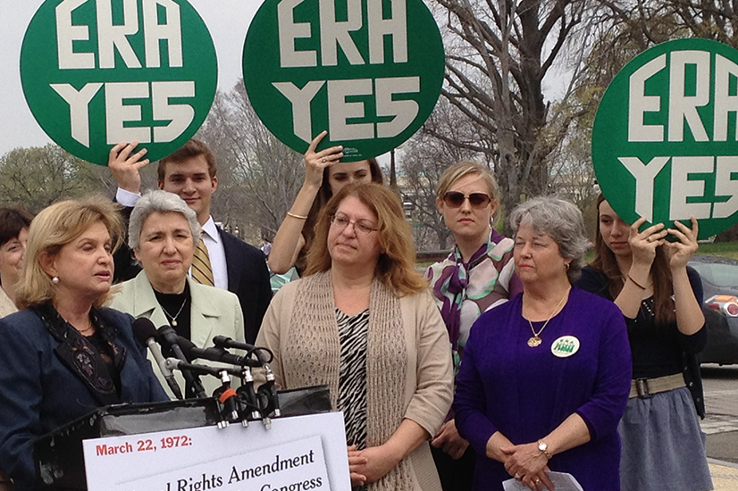 A group of people standing together, with some holding green circular signs reading "ERA YES" in the background. A woman in a navy blue blazer speaks at a podium with multiple microphones.