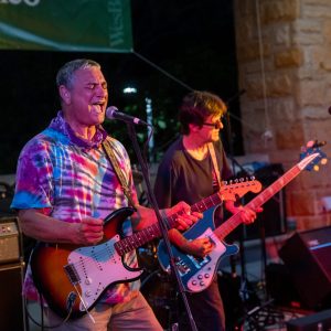 two men standing in the foreground with guitars, playing music and singing into microphones