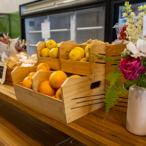 Wooden crates filled with oranges and lemons are displayed on a wooden counter with a vase filled with flowers in shades of pink and green ferns.