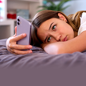 A young woman with long brown hair lies on her bed, resting her head on her arm while holding a smartphone in her hand with a sad expression on her face as she stares at the screen. She is wearing a white t-shirt, and the background features a softly lit bedroom with shelves and a plant.
