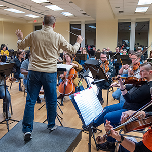 A conductor in a beige sweater and jeans stands on a podium, leading an orchestra rehearsal. The musicians, seated in a semi-circle, play various string and wind instruments while following sheet music in the background.