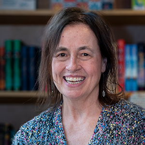 A woman with shoulder-length brown hair wearing a colorful speckled sweater smiles while standing in front of a blurred bookshelf filled books.