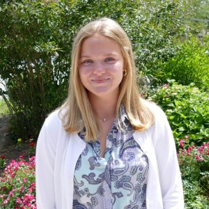 young woman with blonde har, white cardigan and blue floral shirt standing in front of pink flowers outside a library