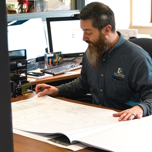 a man with dark hair and a beard sitting at a desk looking over blueprints
