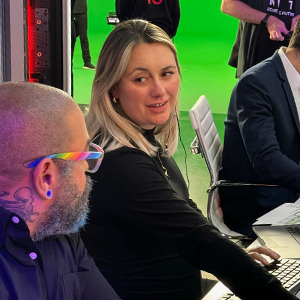 A woman with shoulder length blond hair wearing a black long sleeve top, sitting at a news desk looking towards a man to her left with a bald head and a full beard, wearing colorful glasses and a dark blue button up shirt. Two male colleagues are seated to her right working on computers and camera men stand in front of a green screen in the background.