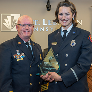 Two uniformed firefighters standing together and smiling, the one on the right is holding a glass "Firefighter of the Year" award. The background is a beige wall with a metal version of the Mt. Lebanon logo.