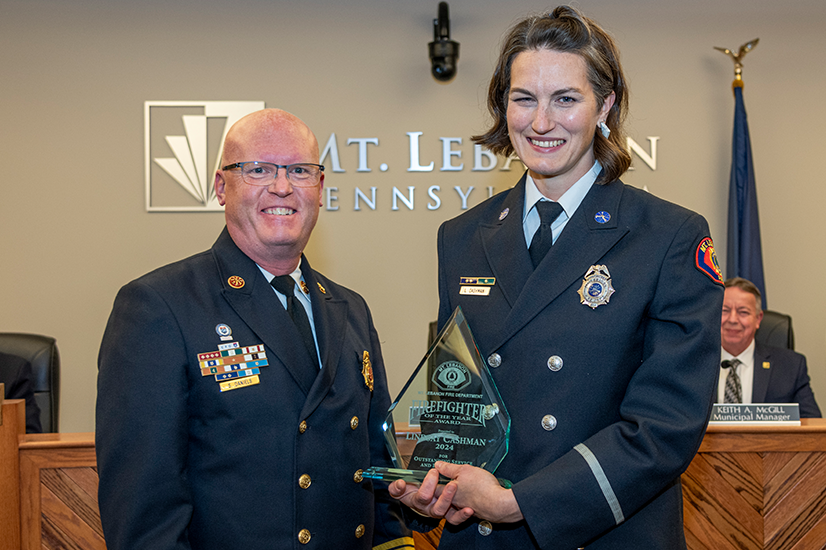 Two uniformed firefighters standing together and smiling, the one on the right is holding a glass "Firefighter of the Year" award. The background is a beige wall with a metal version of the Mt. Lebanon logo.
