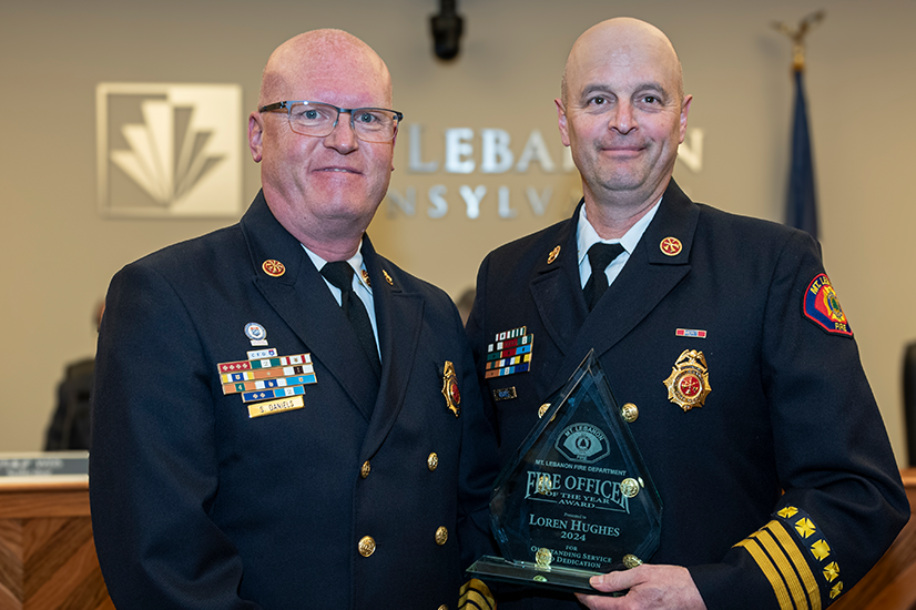 Two uniformed firefighters standing together and smiling, the one on the right is holding a glass "Fire Officer of the Year" award. The background is a beige wall with a metal version of the Mt. Lebanon logo.